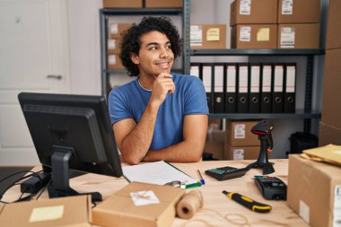 Hispanic man with curly hair working at small business ecommerce with hand on chin thinking about question, pensive expression. smiling and thoughtful face. doubt concept. 