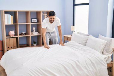 Young hispanic man make bed standing at bedroom