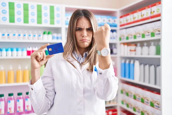 Hispanic young woman working at pharmacy drugstore holding credit card annoyed and frustrated shouting with anger, yelling crazy with anger and hand raised