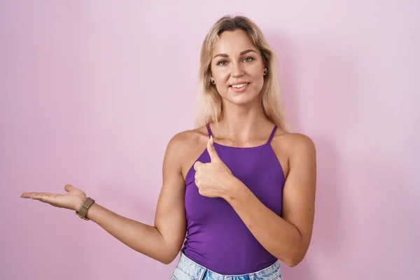Young blonde woman standing over pink background showing palm hand and doing ok gesture with thumbs up, smiling happy and cheerful