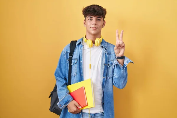 Hispanic teenager wearing student backpack and holding books smiling with happy face winking at the camera doing victory sign with fingers. number two.