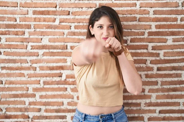Young brunette woman standing over bricks wall punching fist to fight, aggressive and angry attack, threat and violence