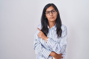 Young hispanic woman wearing glasses pointing with hand finger to the side showing advertisement, serious and calm face 