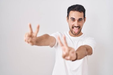 Handsome hispanic man standing over white background smiling with tongue out showing fingers of both hands doing victory sign. number two. 