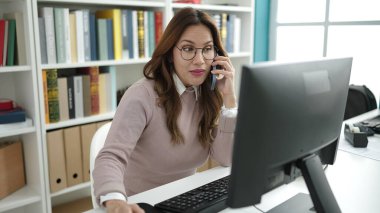 Young beautiful hispanic woman student using computer talking on smartphone at library university