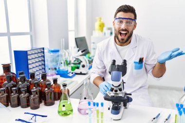 Handsome hispanic man working at scientist laboratory celebrating achievement with happy smile and winner expression with raised hand 