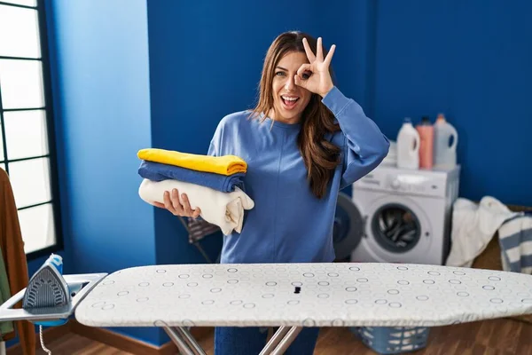Young brunette woman holding folded laundry after ironing smiling happy doing ok sign with hand on eye looking through fingers