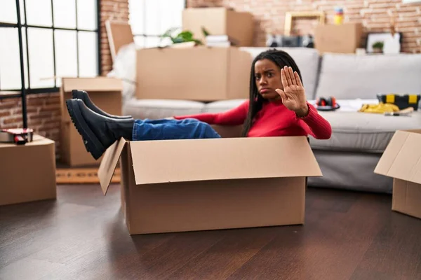 Young african american with braids moving to a new home inside of a cardboard box with open hand doing stop sign with serious and confident expression, defense gesture