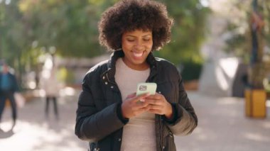 Genç Afrikalı Amerikalı kadın parkta akıllı telefon kullanarak gülümsüyor.