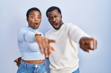 Genç Afro-Amerikan çifti mavi arka planda dikiliyor. Parmakları önde, ağzı açık, şaşkınlık içinde bir ifade sergiliyorlar. 