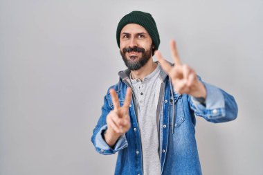 Yün şapkalı, gülümseyen ve parmaklarını zafer işareti yaparken gösteren İspanyol dövmeli genç adam. İki numara.. 