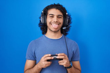 Hispanic man with curly hair playing video game holding controller sticking tongue out happy with funny expression. 