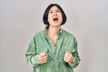 Young asian woman standing over white background angry and mad screaming frustrated and furious, shouting with anger. rage and aggressive concept. 