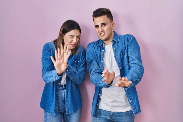 Young hispanic couple standing over pink background disgusted expression, displeased and fearful doing disgust face because aversion reaction.