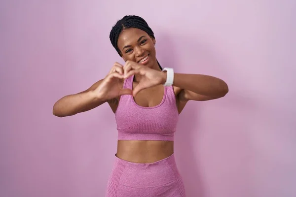 戴着辫子 身穿运动服 背景粉红的非洲裔美国女人 带着爱的笑容用手做着心形符号 浪漫的概念 — 图库照片