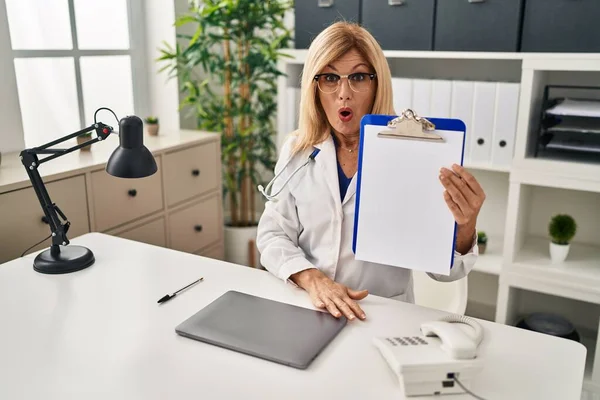 Middle Age Blonde Doctor Woman Holding Clipboard Clinic Scared Amazed — Stockfoto