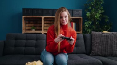 Young blonde woman playing video game sitting on sofa at home