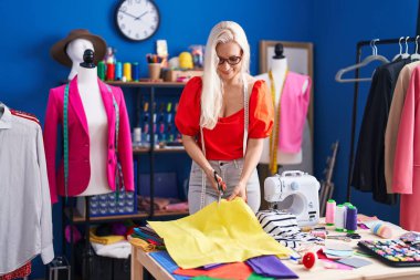 Young blonde woman tailor smiling confident cutting cloth at sewing studio