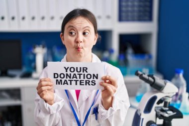 Woman with down syndrome working at scientist laboratory holding your donation matters banner making fish face with mouth and squinting eyes, crazy and comical. 