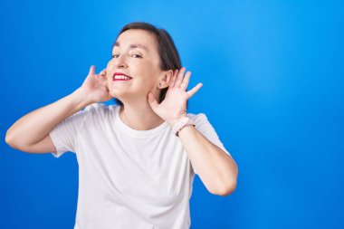 Middle age hispanic woman standing over blue background trying to hear both hands on ear gesture, curious for gossip. hearing problem, deaf 