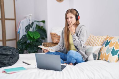 Young caucasian woman studying with laptop sitting on the bed covering mouth with hand, shocked and afraid for mistake. surprised expression 