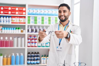 Young arab man pharmacist smiling confident holding glasses at pharmacy