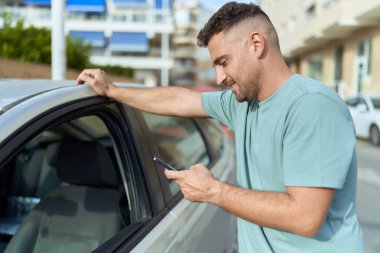 Young hispanic man using smartphone leaning on car at street