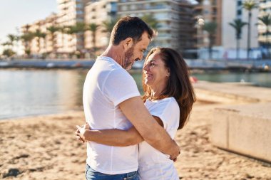 Orta yaşlı erkek ve kadın çift sahilde birbirlerine sarılıyorlar.