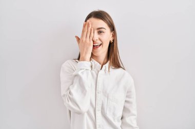 Young caucasian woman standing over isolated background covering one eye with hand, confident smile on face and surprise emotion. 