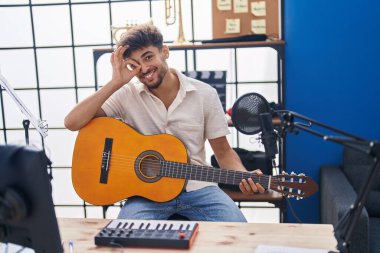 Arab man with beard playing classic guitar at music studio smiling happy doing ok sign with hand on eye looking through fingers 