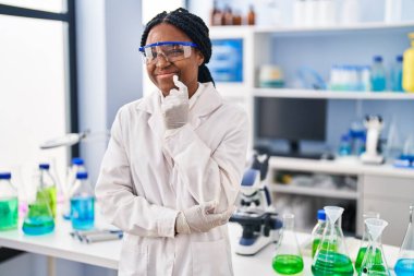 African american woman working at scientist laboratory thinking concentrated about doubt with finger on chin and looking up wondering 