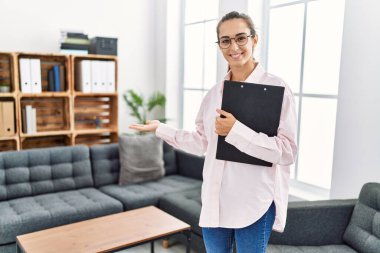 Young hispanic woman smiling confident holding clipboard standing at clinic