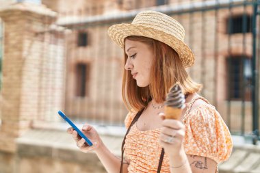 Genç kızıl saçlı turist akıllı telefon kullanıyor ve sokakta dondurma yiyor.