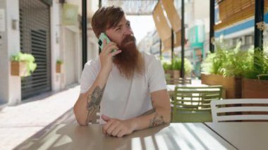 Young redhead man talking on the smartphone sitting on table at coffee shop terrace