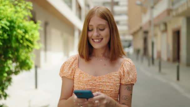 红头发的年轻女子自信地微笑着 用智能手机在街上走着 — 图库视频影像