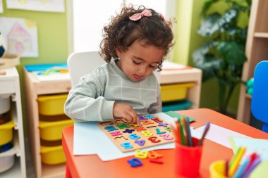 Adorable hispanic girl playing with maths puzzle game sitting on table at kindergarten