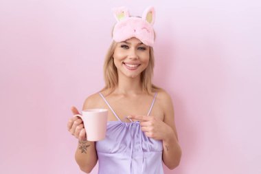 Young caucasian woman wearing sleep mask drinking a cup of coffee smiling happy pointing with hand and finger 