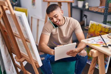 Young hispanic man artist talking on the smartphone drawing on notebook at art studio