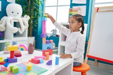 Adorable hispanic boy playing with construction blocks standing at kindergarten