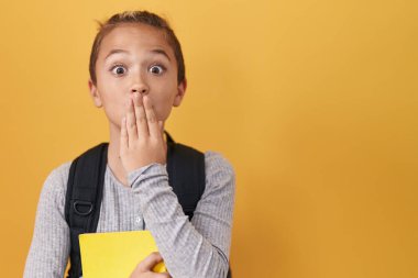 Little caucasian boy wearing student backpack and holding book covering mouth with hand, shocked and afraid for mistake. surprised expression 