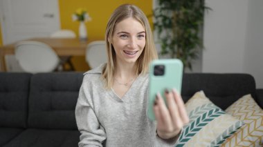 Young blonde woman taking selfie picture with smartphone sitting on the sofa at home