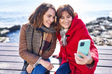 Two women mother and daughter make selfie by smartphone sitting on bench at seaside