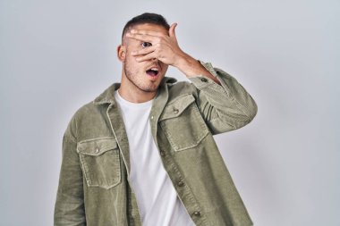 İzole edilmiş arka planda duran genç İspanyol adam şok içinde yüzünü ve gözlerini kapayarak utangaç ifadesiyle parmaklarının arasından bakıyor.. 