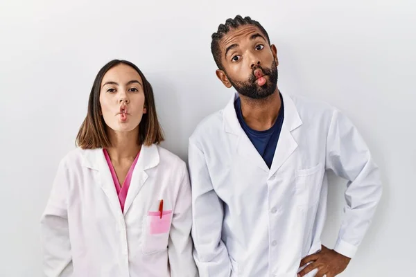 Young hispanic doctors standing over white background making fish face with lips, crazy and comical gesture. funny expression.