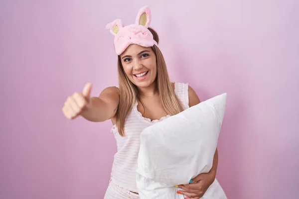 Young blonde woman wearing pyjama hugging pillow approving doing positive gesture with hand, thumbs up smiling and happy for success. winner gesture.