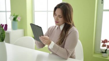 Genç ve güzel bir kadın günlük giysiler içinde kameraya bakıyor ve gülümsüyor, bir koltukta oturuyor ve tablet kullanıyor.