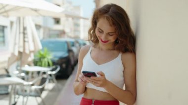 Genç, güzel, İspanyol bir kadın sokakta akıllı telefon kullanarak gülümsüyor.