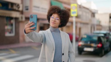 Afro-Amerikalı kadın gülümsüyor. Sokakta akıllı telefonun yanında özçekim yapıyor.