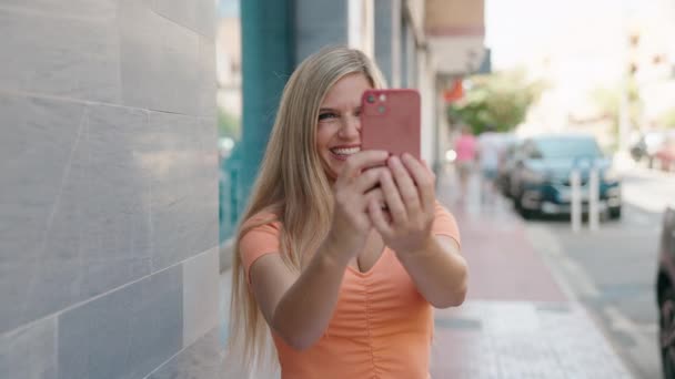금발의 여성은 자신감을 가지고 웃으면서 거리에서 스마트폰으로 사진을 찍는다 — 비디오