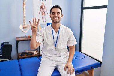 Young hispanic man with beard working at pain recovery clinic showing and pointing up with fingers number four while smiling confident and happy. 